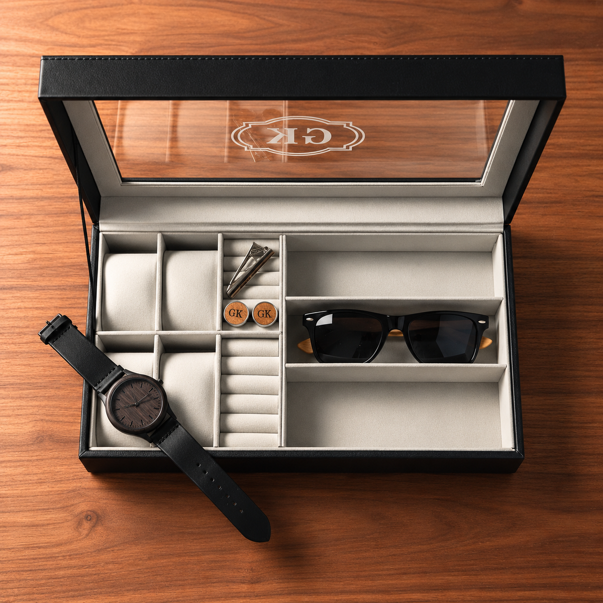 An open black watch box with a clear lid, displayed on a wooden surface. The box contains multiple compartments, including a watch slot, ring holder, coin compartment, and a sunglasses slot.