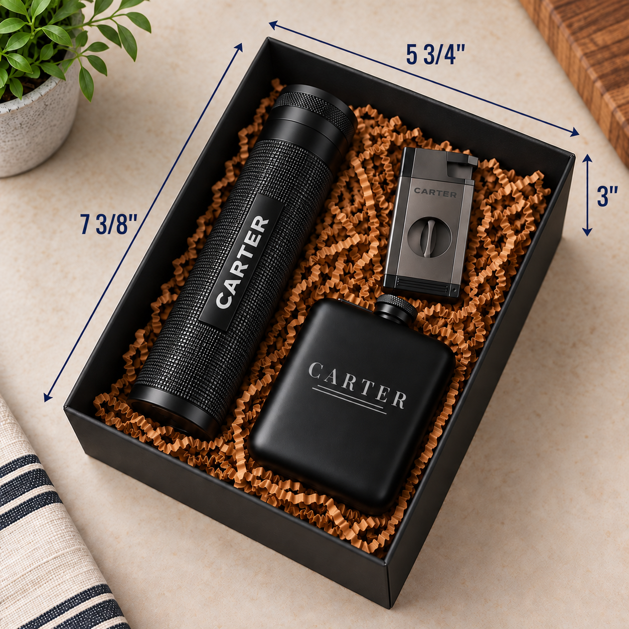 A black gift box filled with brown crinkle paper packaging holds a black cylindrical bottle labeled CARTER, a black rectangular lighter, and a black flask labeled CARTER. Blue dimension arrows and text show the box measurements: 7 3/8 inches tall, 5 3/4 inches wide, and 3 inches deep.