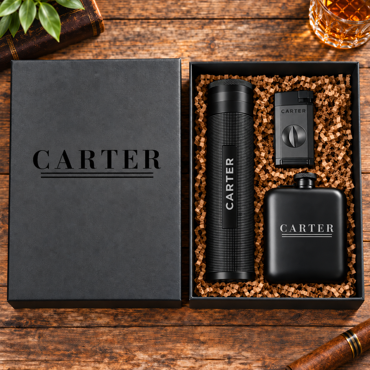 A luxury gift set from CARTER, featuring a matte black metal flask, a matching cylindrical lighter, and a small rectangular lighter, all resting in a black presentation box filled with brown crinkle paper. The black lid of the box is shown next to it on a rustic wooden table, with a cigar, a glass of whiskey, and green leaves as styled props.