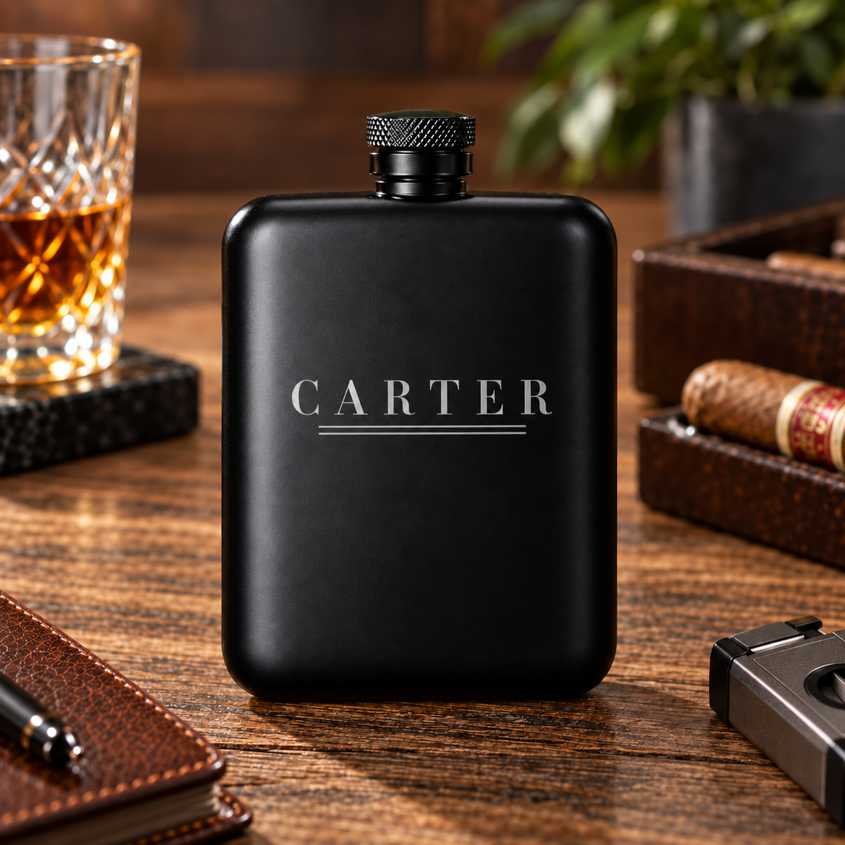 A matte black metal flask with the name 'CARTER' in silver lettering, set on a wooden table surrounded by lifestyle props including a glass of whiskey, a cigar, a notebook, and a cigar box.