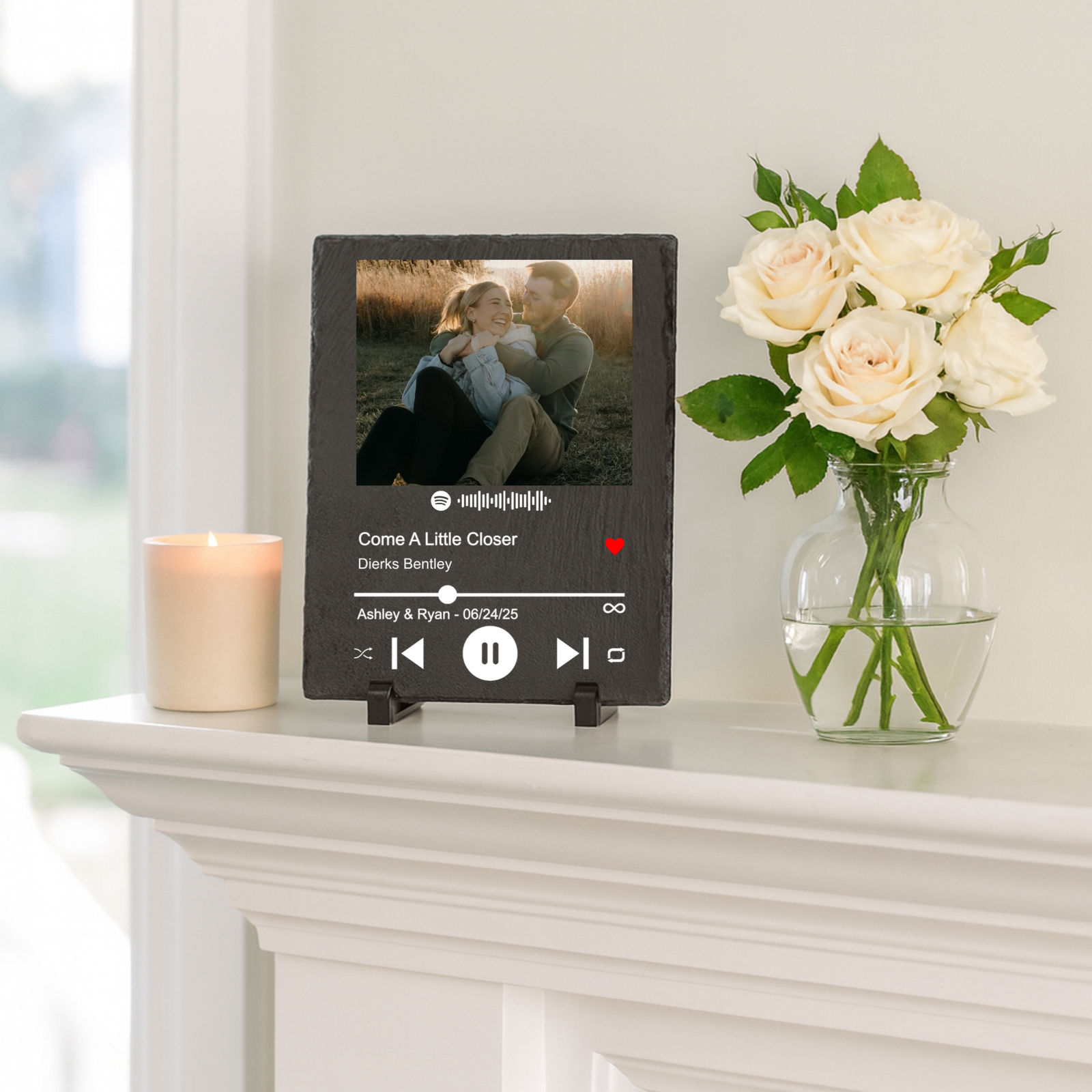 Slate plaque personalized with sample photo and Spotify player interface displaying song "Come A Little Closer" by Dierks Bentley with names Ashley & Ryan and date 06/24/25, displayed on mantle with candle and flowers.