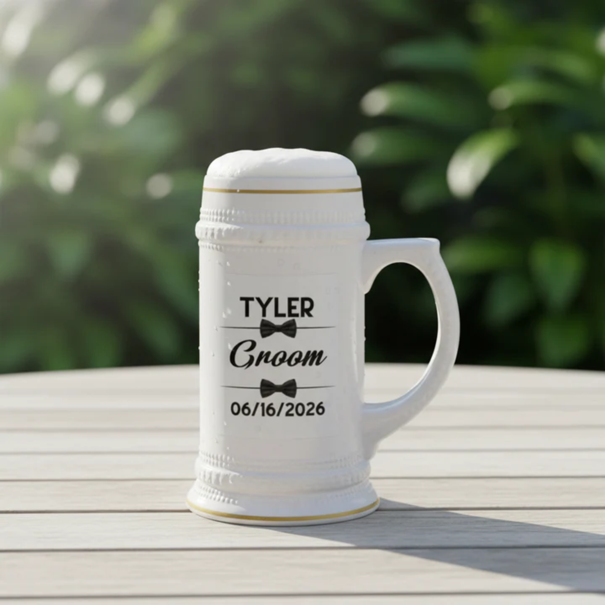 White ceramic beer stein with sample engraving showing name "TYLER," text "Groom," bow tie graphics, and sample date "06/16/2026," displayed on wooden surface with blurred green foliage background.