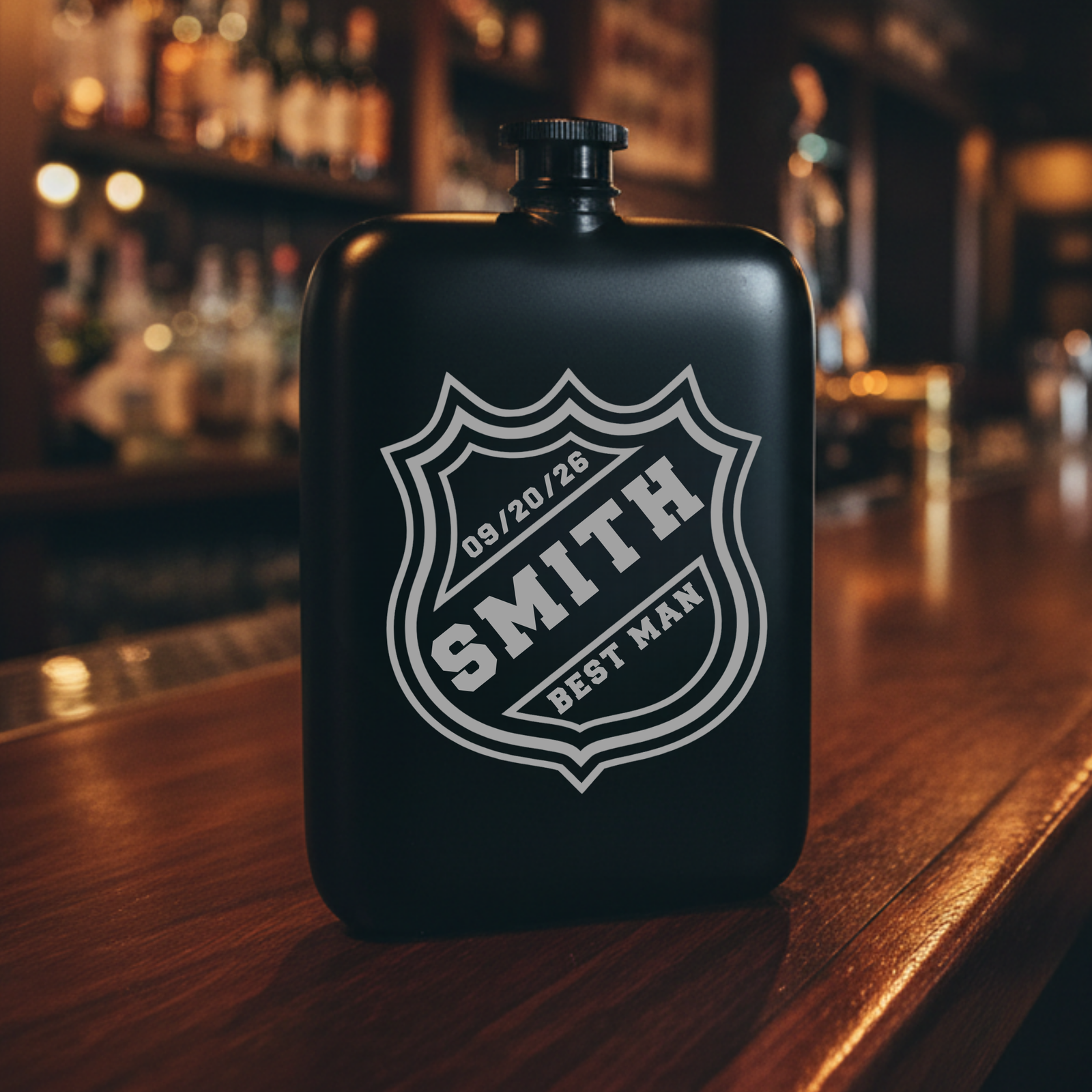 Black stainless steel flask with laser-engraved hockey crest shield displaying sample personalization "SMITH," "03/20/26," and "BEST MAN" text, shown on wooden bar counter with blurred bar background.