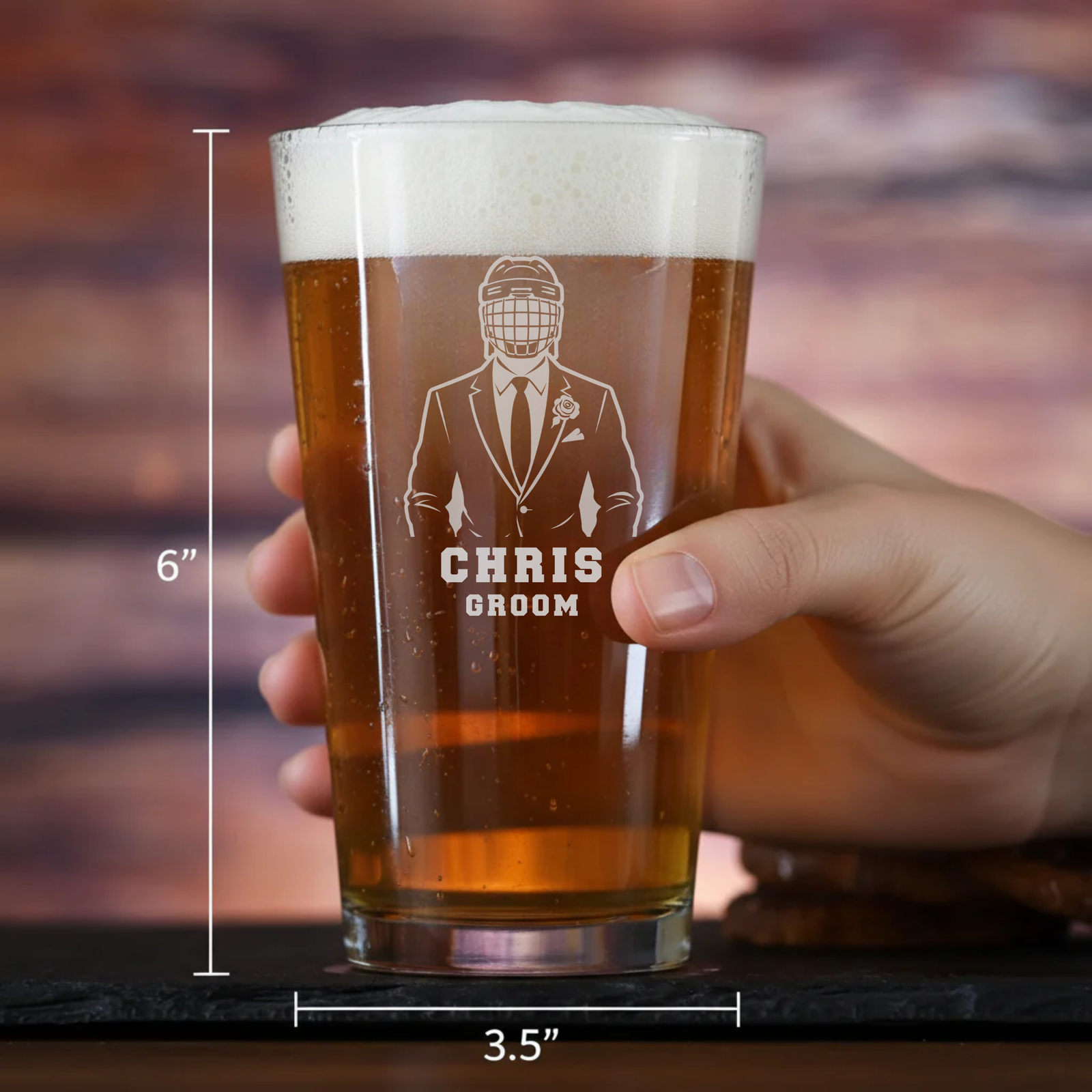 Pint glass filled with beer, laser-engraved with sample design showing groomsman in hockey helmet and "CHRIS GROOM" text, held in hand with 6" height and 3.5" width dimensions marked