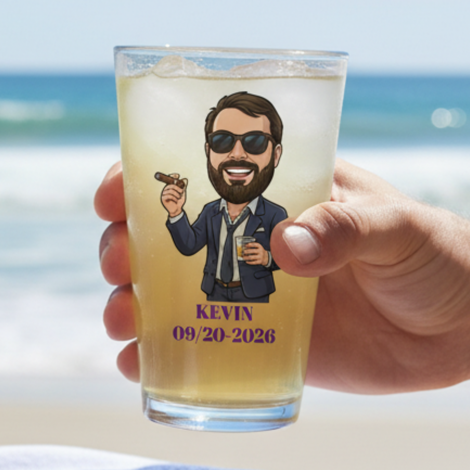 Hand holding pint glass filled with pale beverage, printed with sample caricature of man in sunglasses and blazer holding cigar, personalized with name "Kevin" and date "09/20-2026" in purple text, ocean beach backdrop.