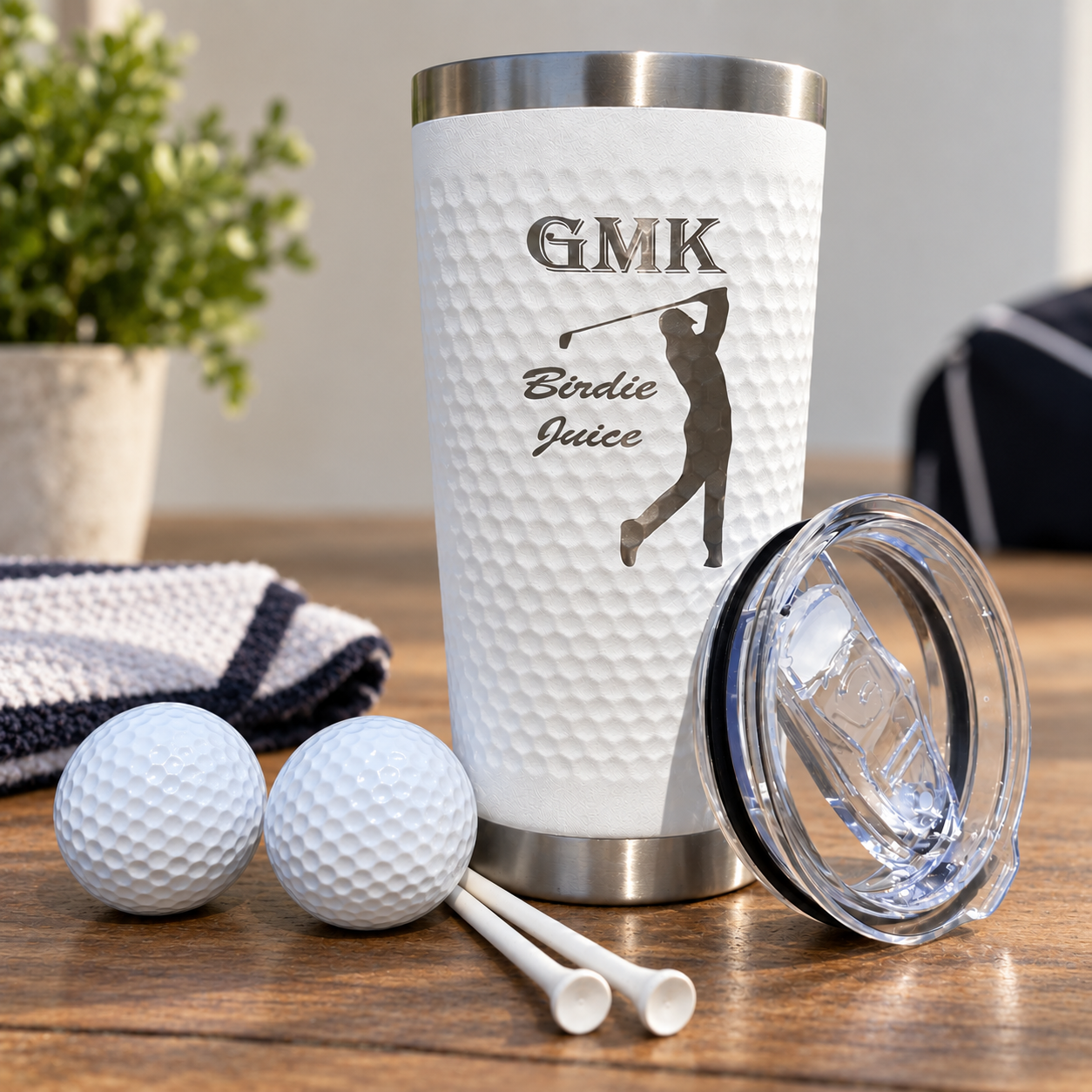 A white textured insulated tumbler with a stainless steel lid sits on a wooden table, shown with two golf balls, two tees, and a clear lid placed beside it. The tumbler is printed with the text 'GMK Birdie Juice' and a golfer silhouette.