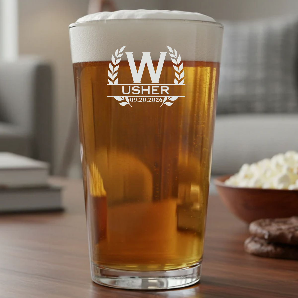 Pint glass filled with beer, laser-engraved with sample design showing "W" initial, "USHER" text, and date "09.20.2026" with decorative laurel wreath motif