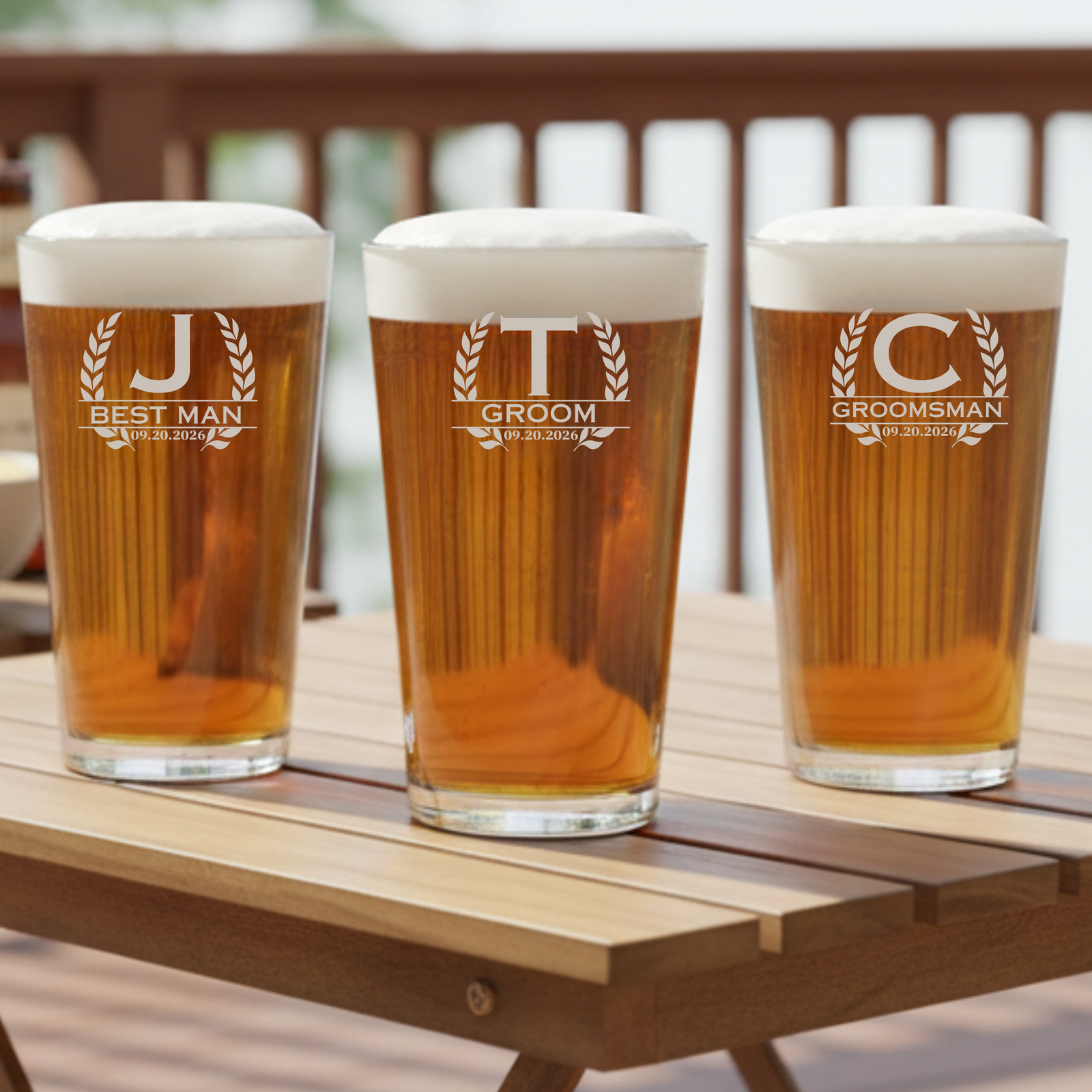 Three personalized pint glasses engraved with sample initials J, T, and C with laurel wreath borders and date 09.20.2026, filled with beer on wooden serving board