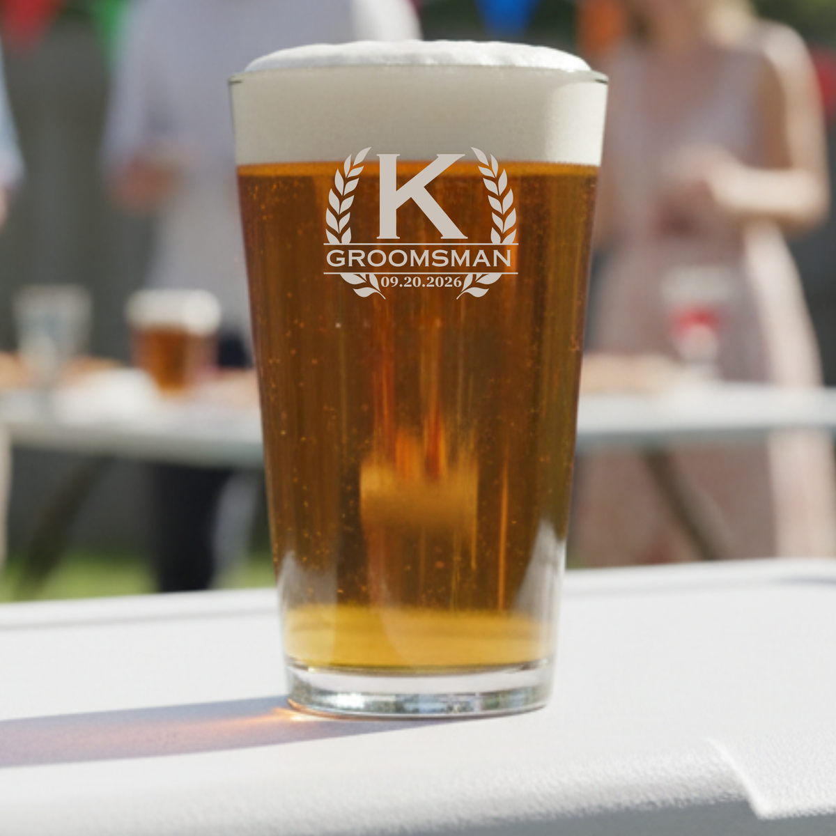 Pint glass filled with beer, etched with sample monogram "K" flanked by laurel wreaths and text "GROOMSMAN 09.20.2026
