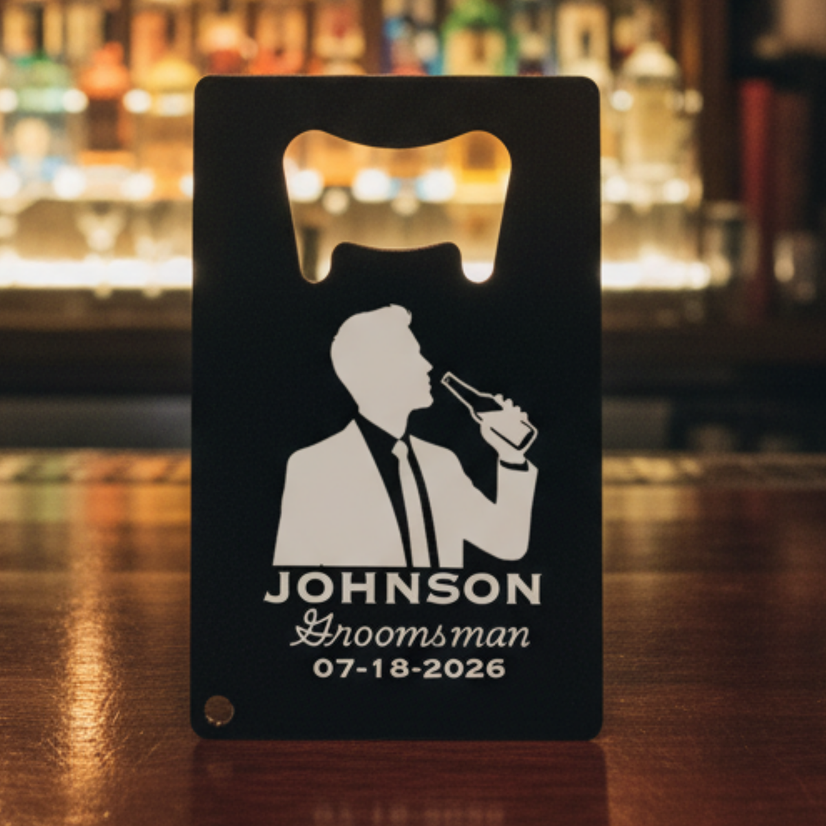 Black stainless steel card opener with laser-engraved silhouette of man in tuxedo holding beer, personalized with sample name "JOHNSON," title "Groomsman," and date "07-18-2026
