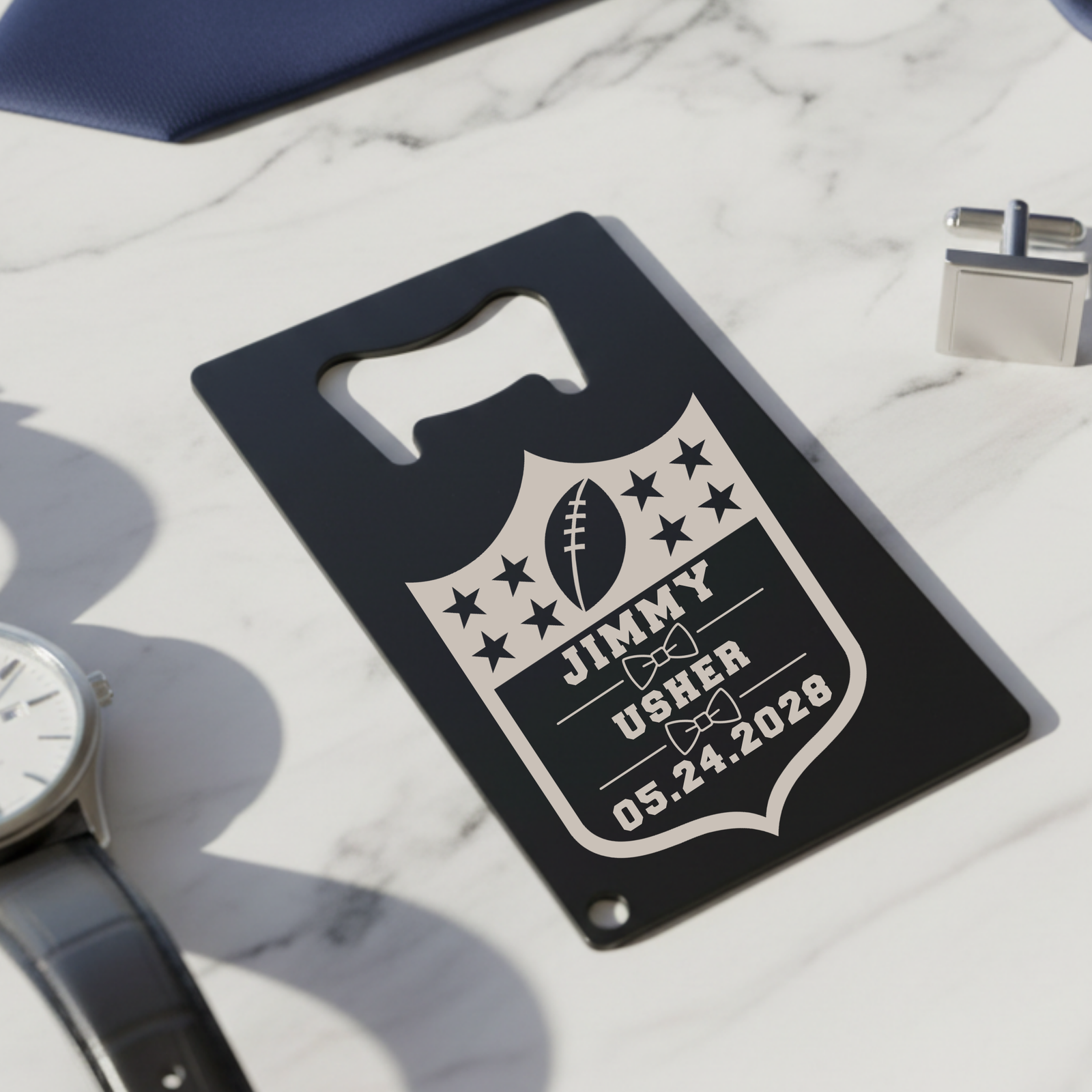 Black stainless steel bottle opener with engraved football shield design featuring sample personalization "JIMMY USHER 05.24.2028" and star motifs, displayed on marble surface with watch and cufflinks.