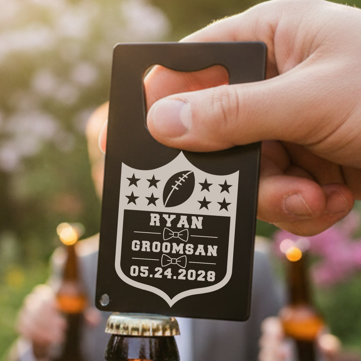 Black stainless steel bottle opener with shield design, personalized with sample name "Ryan," football motif, bow-tie icons, and wedding date "05.24.2028," held in hand outdoors.