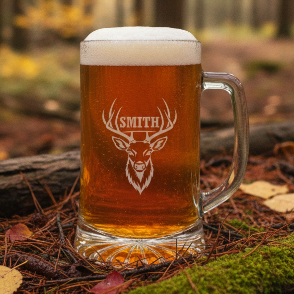 Engraved glass beer mug filled with amber beer and foam, featuring a sample personalization with a deer head design and the name "SMITH" in a forest setting.