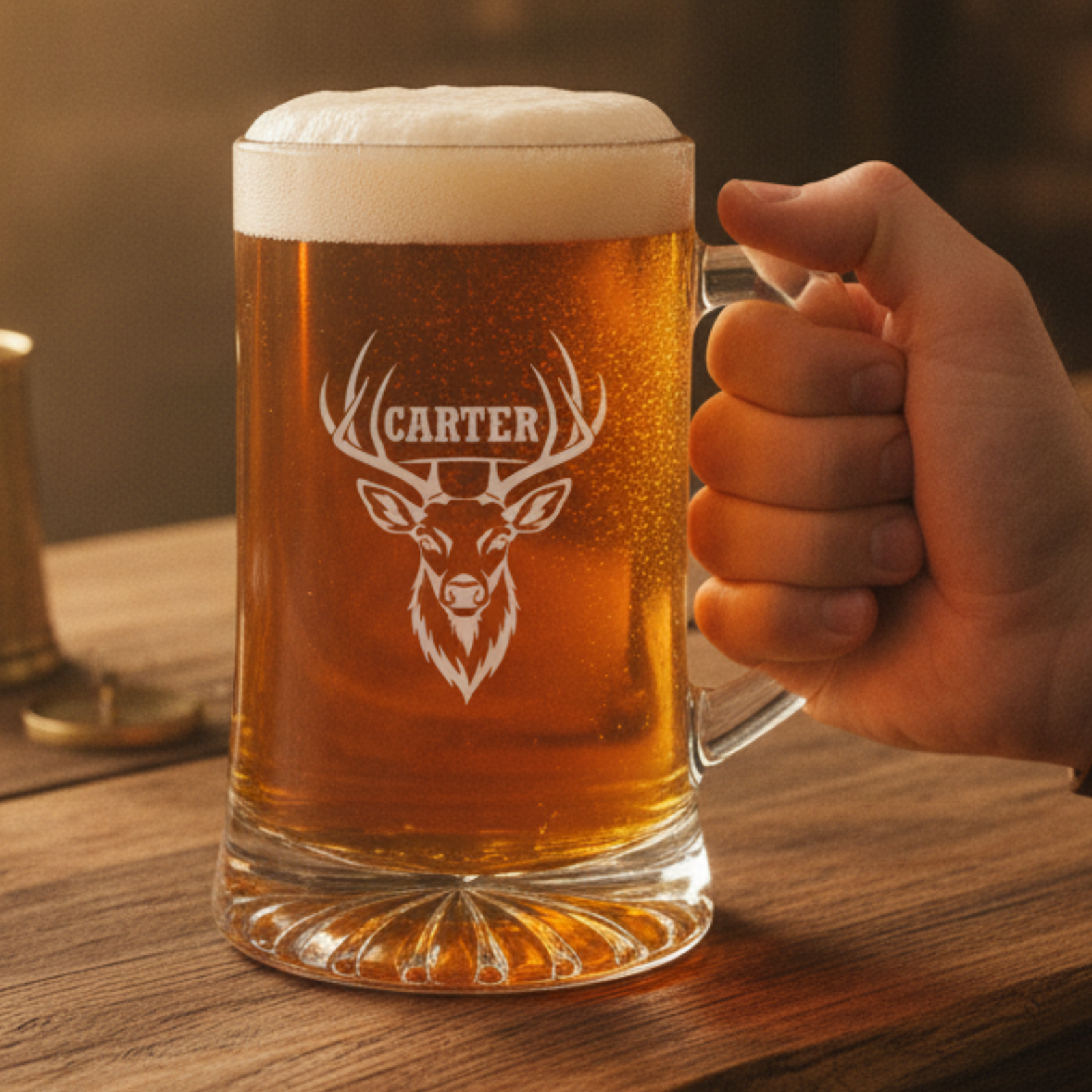Hand holding engraved beer mug with deer head and "CARTER" name sample, filled with amber beer and foam head