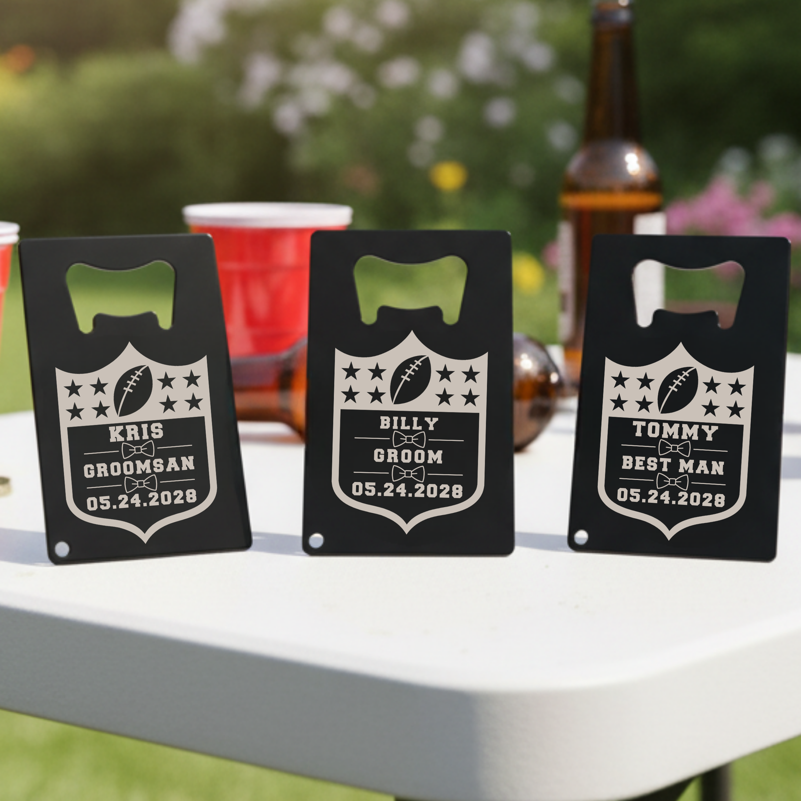 Three black bottle openers with personalized engraving samples reading "KRIS GROOMSMAN," "BILLY GROOM," and "TOMMY BEST MAN" with football shield design and date 05.24.2028, displayed outdoors on white surface