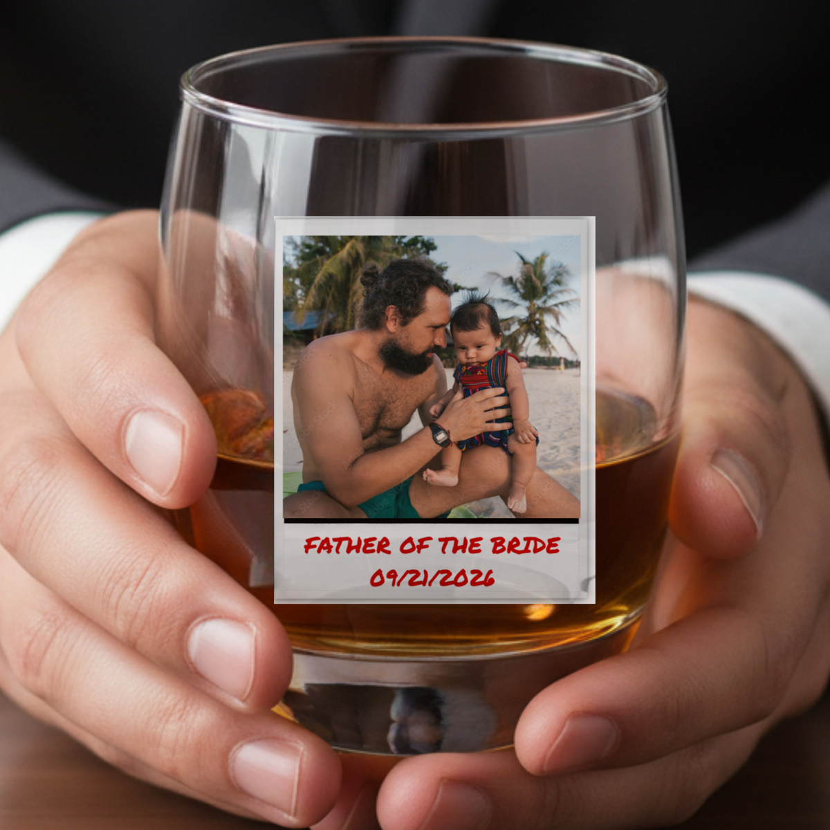 Whiskey glass held in hand, filled with amber liquid, featuring a custom photo printed as a Polaroid-style image with sample text "Father of the Bride" and date "09/21/2026