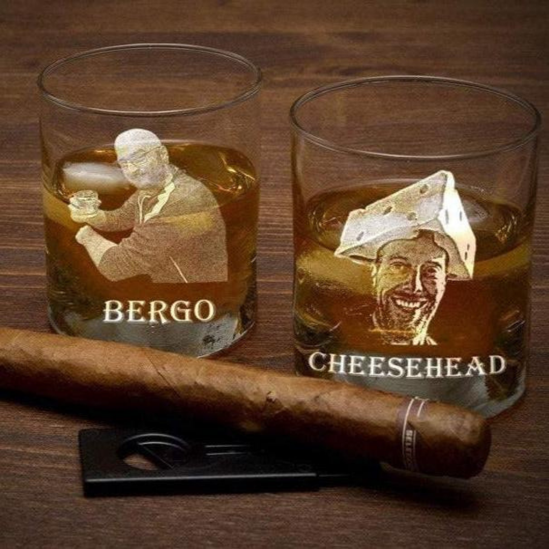 Two laser-engraved whiskey glasses with sample designs featuring portrait images labeled "BERGO" and "CHEESEHEAD," filled with amber liquid and positioned beside a cigar on wooden surface