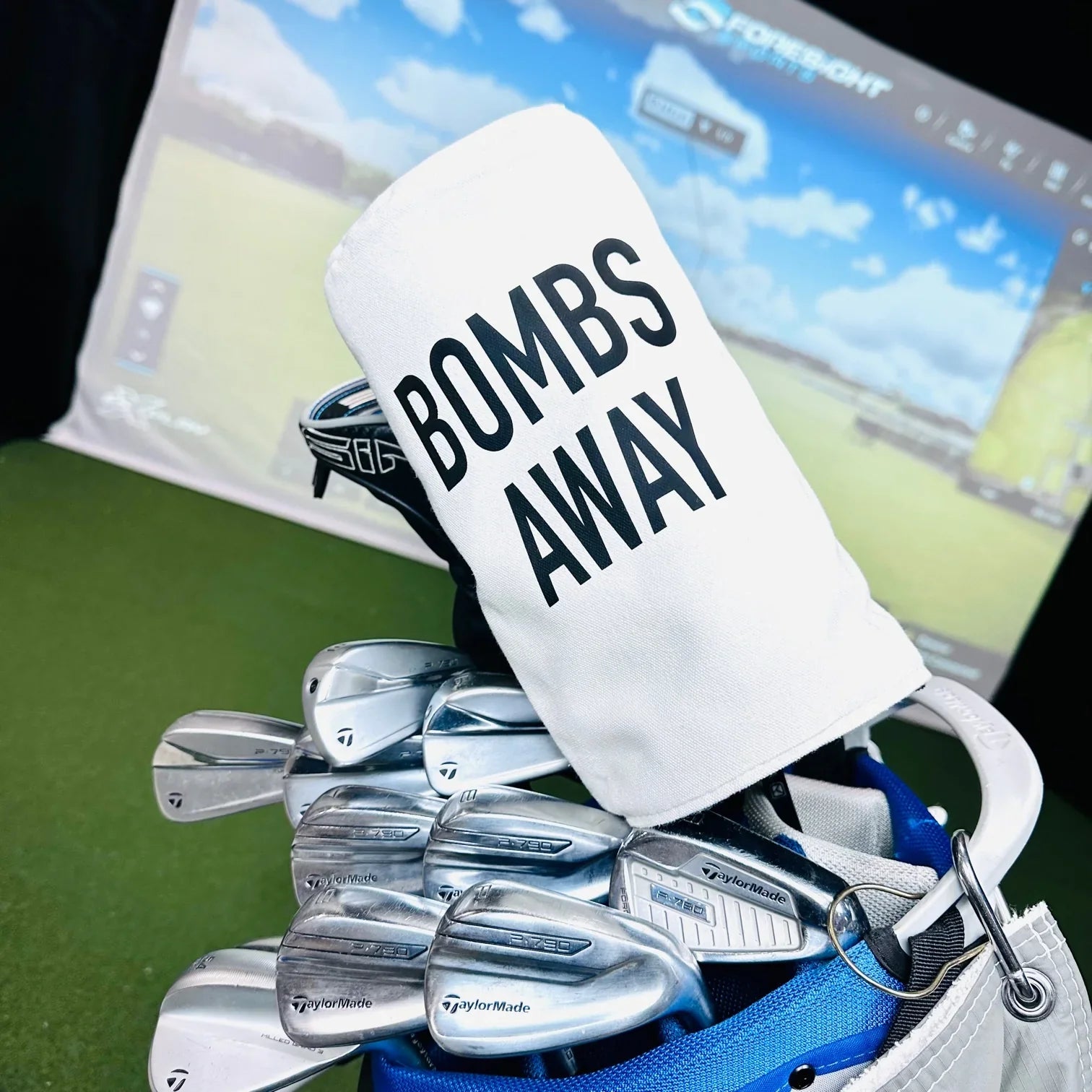 White barrel head cover printed with "BOMBS AWAY" text, displayed on golf clubs in a golf bag with course visible in background