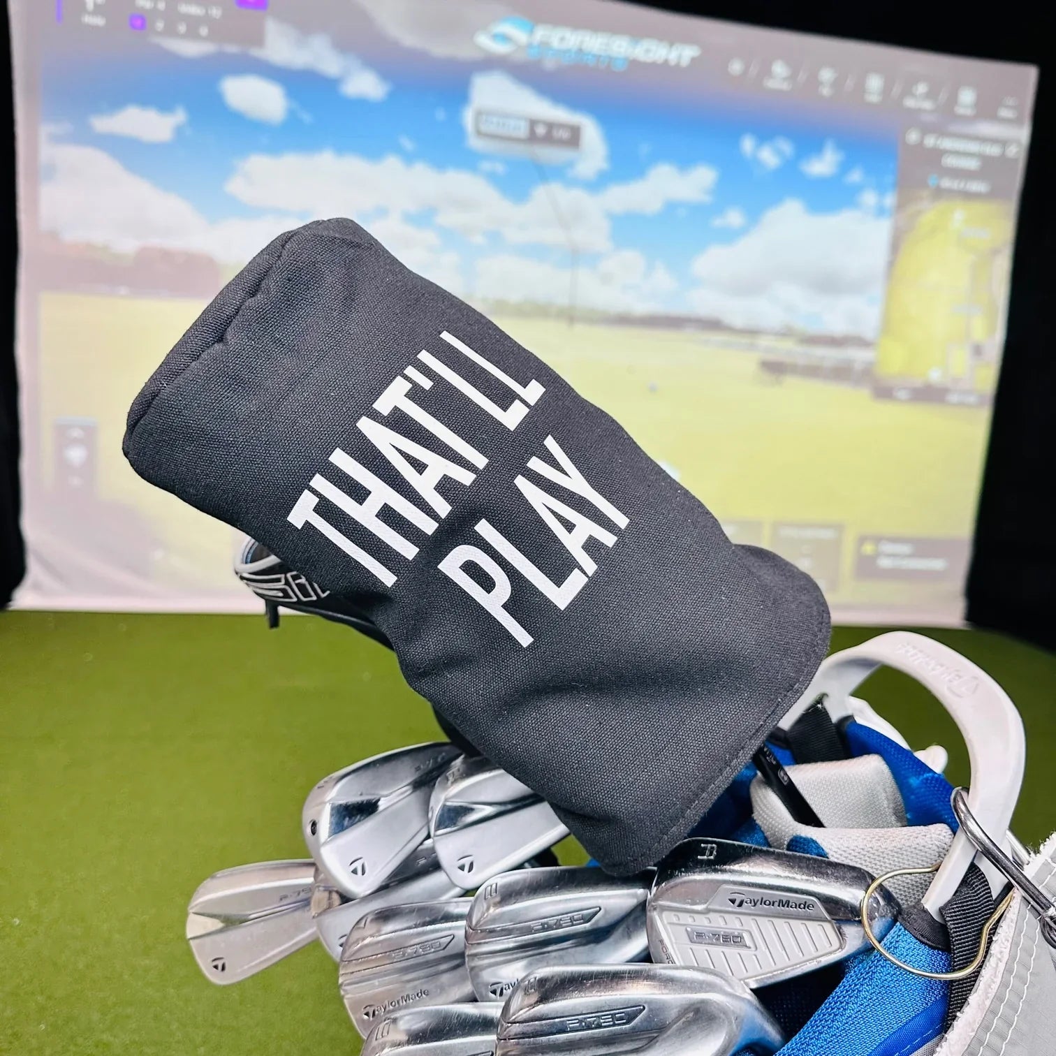 Charcoal gray golf head cover printed with white text reading "THAT PLAY" positioned over golf clubs on grass with simulator screen in background