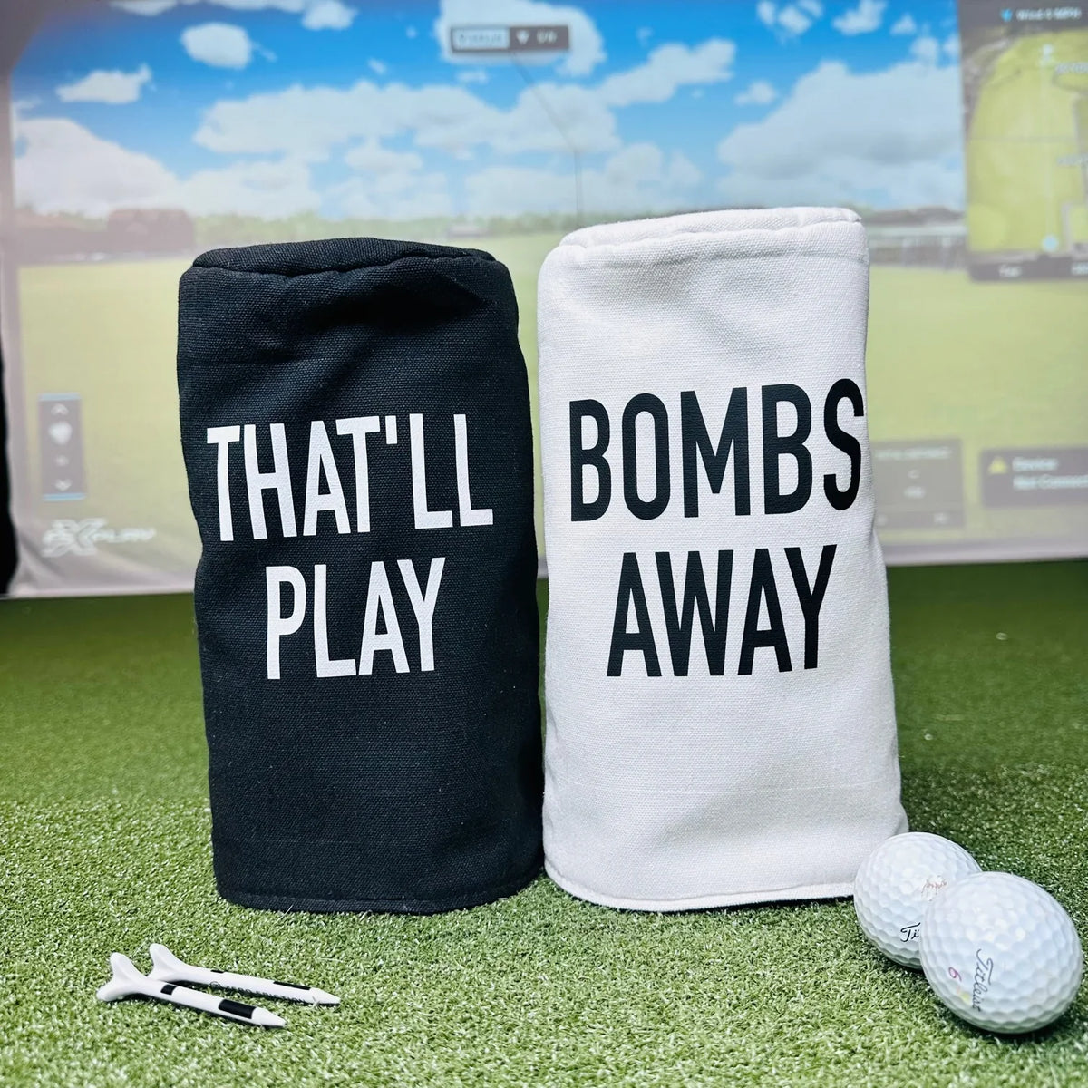 Black and white barrel head covers printed with "THAT'LL PLAY" and "BOMBS AWAY" text, displayed on artificial turf with golf balls and tees