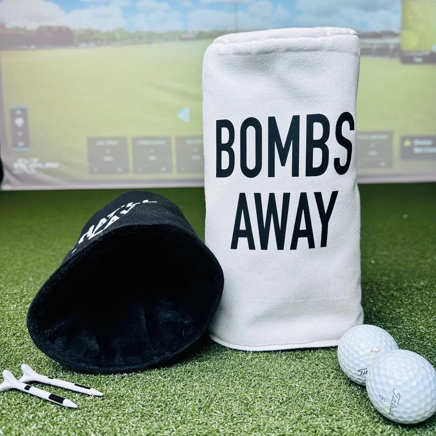 White barrel head cover printed with "BOMBS AWAY" text, displayed with black head cover, golf balls, and tees on artificial turf with stadium background