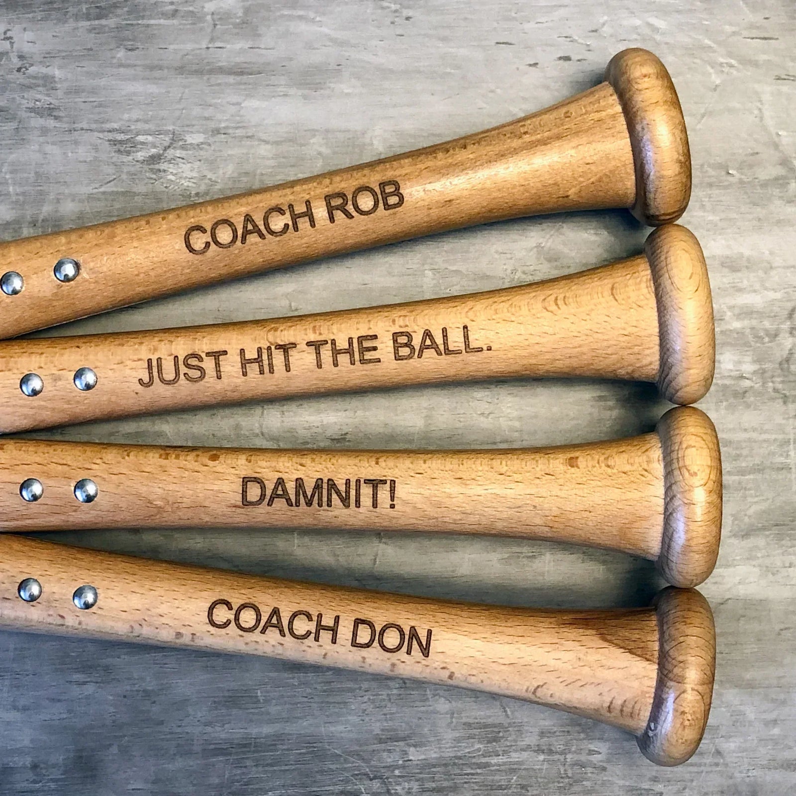 Four wooden baseball bat bottle openers with sample engraved text including "COACH ROB," "JUST HIT THE BALL," "DAMNIT!" and "COACH DON