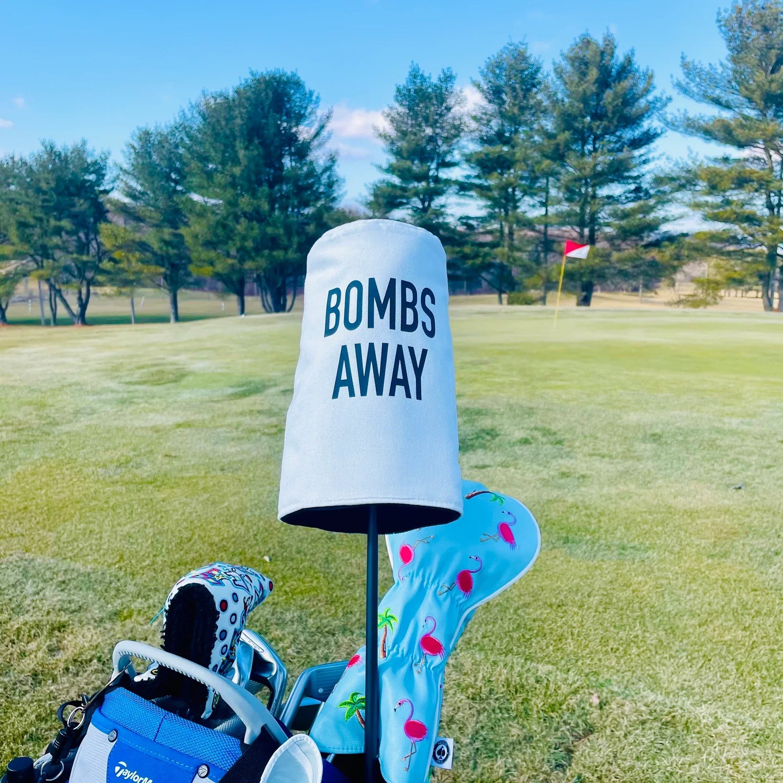Personalized barrel head cover with sample text "BOMBS AWAY" displayed on golf course with patterned club covers in background