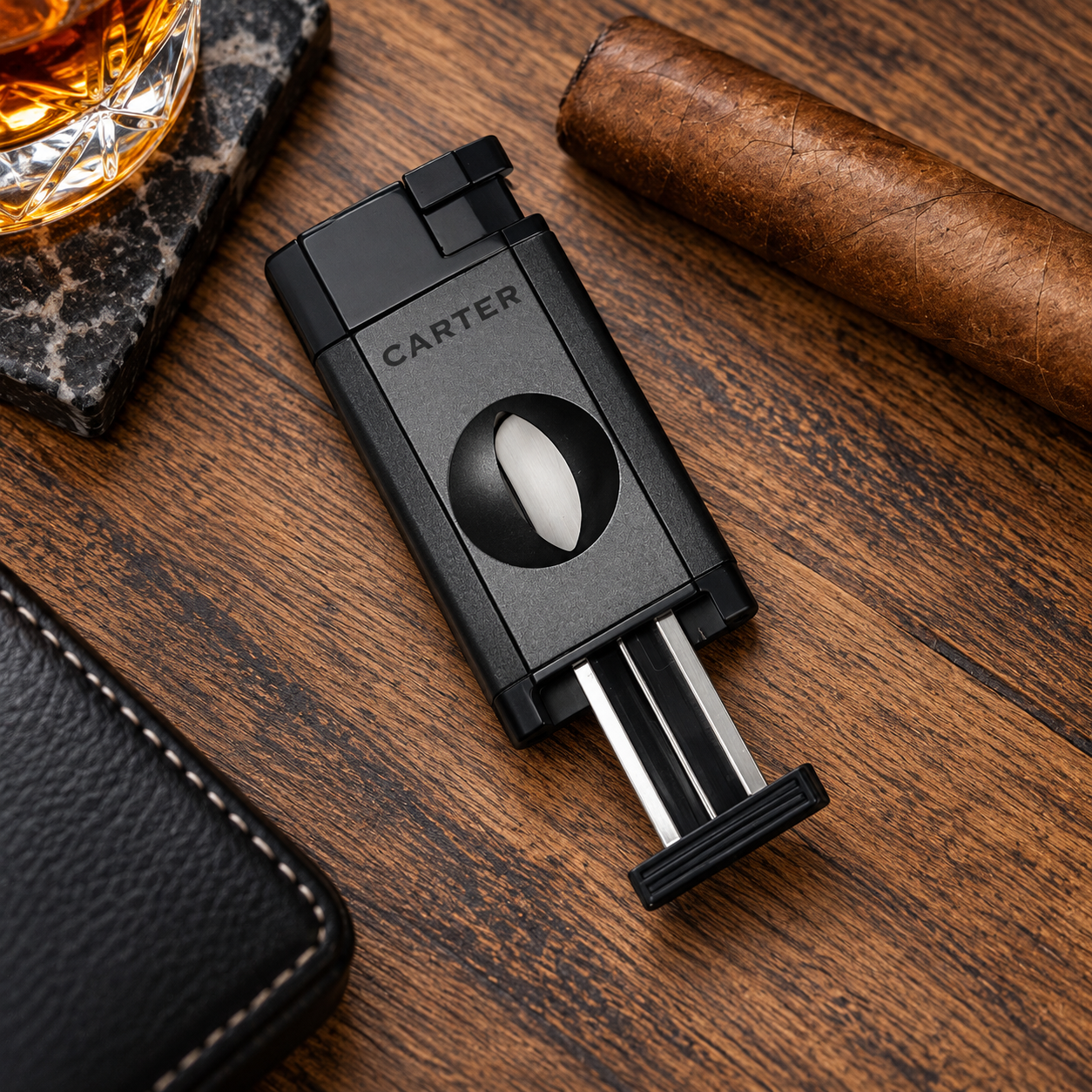 A black Carter cigar humidor with a visible metal wick, photographed on a wooden tabletop alongside a cigar, a glass of amber liquor, and a leather accessory.