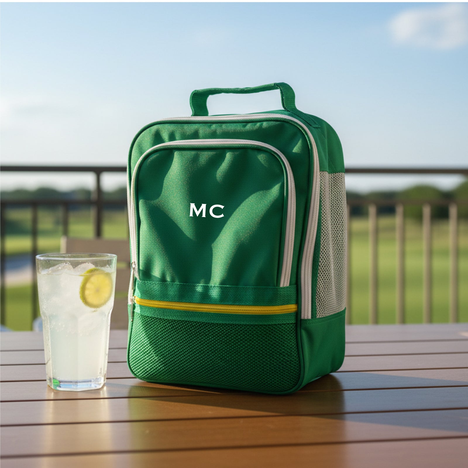 Green shoe bag with monogrammed sample initials "MC" displayed outdoors on wooden deck next to beverage glass