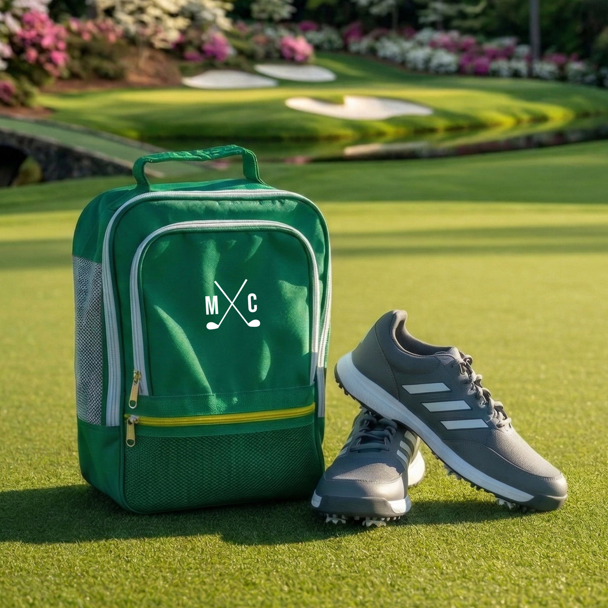 Green golf shoe bag with gray trim and sample monogram "M X C" in white, displayed on putting green next to golf shoes
