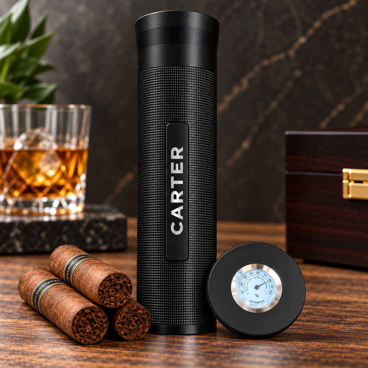 Black textured cylindrical cigar case with sample engraving "CARTER" displayed beside cigars, whiskey glass, thermometer, and dark wooden box