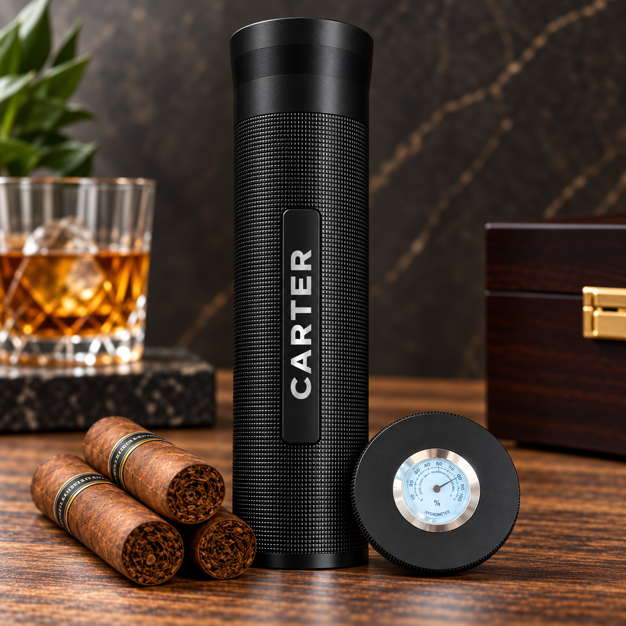 Black textured cylindrical cigar case with sample engraving "CARTER" displayed beside cigars, whiskey glass, thermometer, and dark wooden box