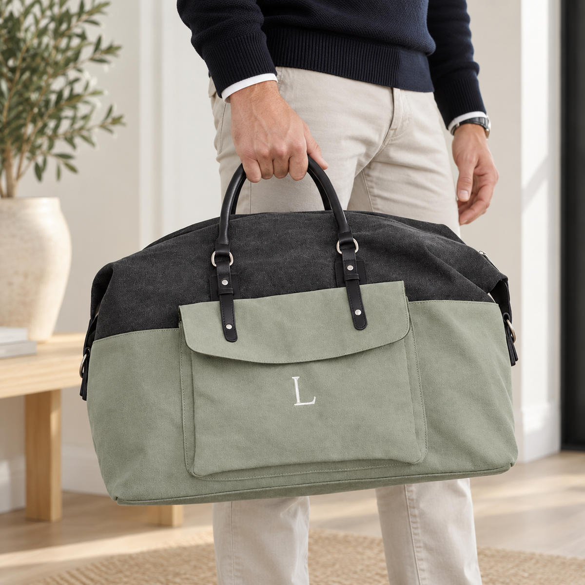 A person in a navy sweater and beige pants holds a large duffel bag with a dark gray top half, sage green bottom half, black handles, and a white embroidered letter 'L' on the front pocket. The setting is a bright indoor room with a wooden table and potted plant in the background.