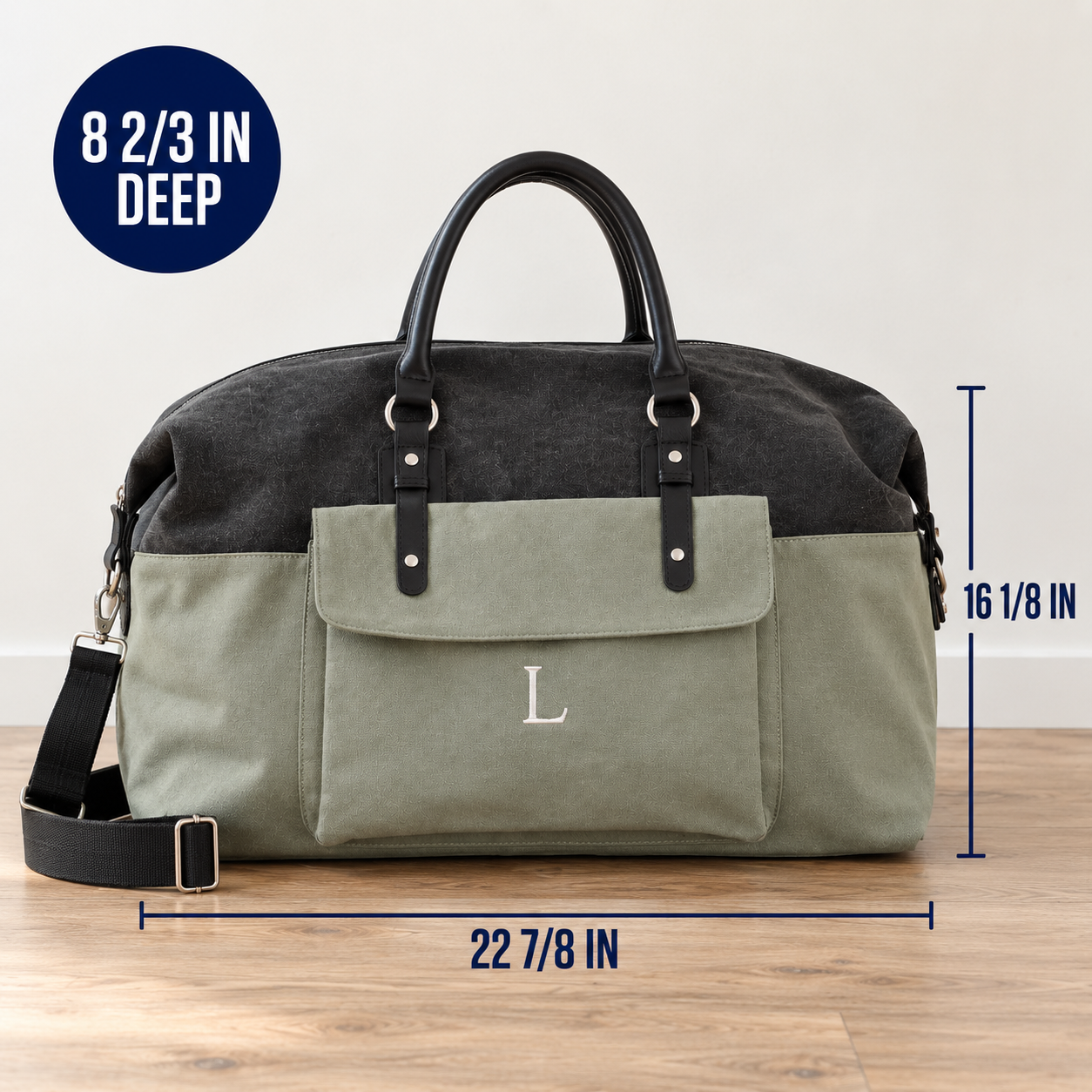 A two-tone duffel bag with a dark charcoal top half and an olive green bottom half, featuring black leather handles and a front pocket with a silver monogram 'L'. The image includes dimension lines showing 22 7/8 inches wide, 16 1/8 inches tall, and an 8 2/3 inch deep capacity, set on a wooden floor against a white wall.