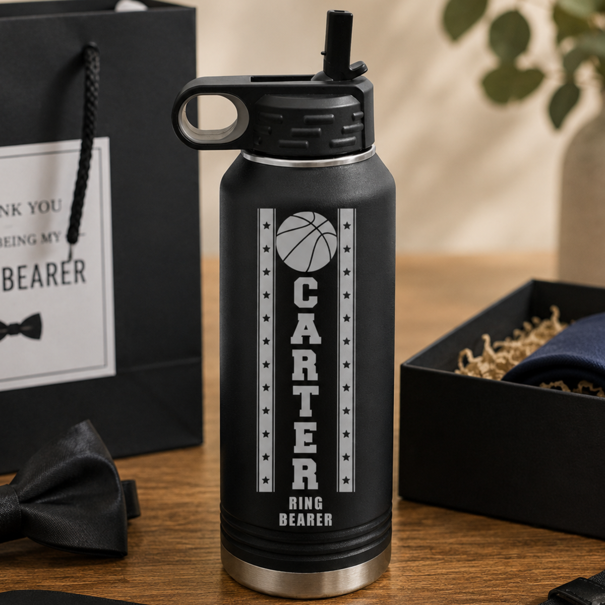 Black insulated water bottle engraved with basketball design and "CARTER RING BEARER" text, displayed with wedding party gifts including ring box and bow tie.