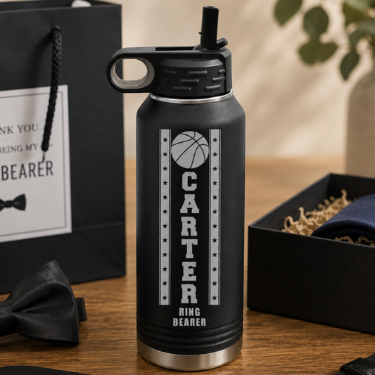 Black insulated water bottle engraved with basketball design and "CARTER RING BEARER" text, displayed with wedding party gifts including ring box and bow tie.
