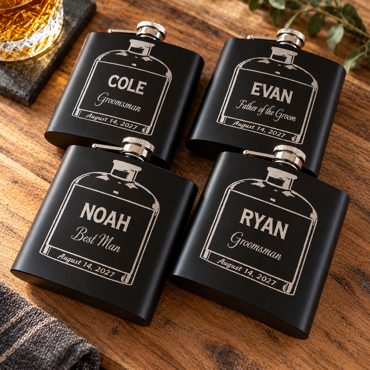 Four black engraved whiskey-style flasks arranged on a wooden surface, each with a bottle-outline label and personalized names: Cole, Evan, Noah, and Ryan, plus wedding/date text. A glass of amber drink and dark stones are partially visible around the scene.