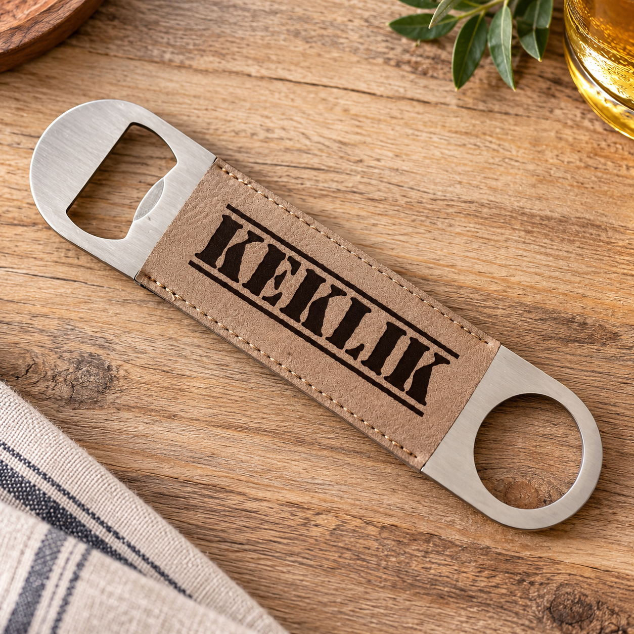 A personalized bottle opener with a tan faux-leather center strap and silver metal ends, engraved with the name 'KEKLIK', shown on a wooden tabletop with a striped cloth and a drink partially visible.