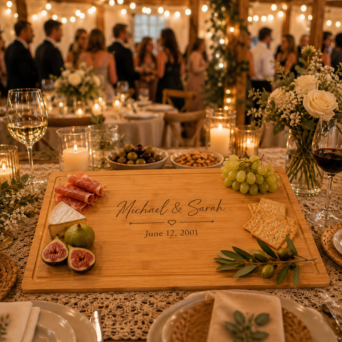 A personalized wooden charcuterie board engraved with 'Michael &amp; Sarah' and a date, styled on a decorated wedding reception table with cheese, grapes, crackers, olives, and candles in the background.