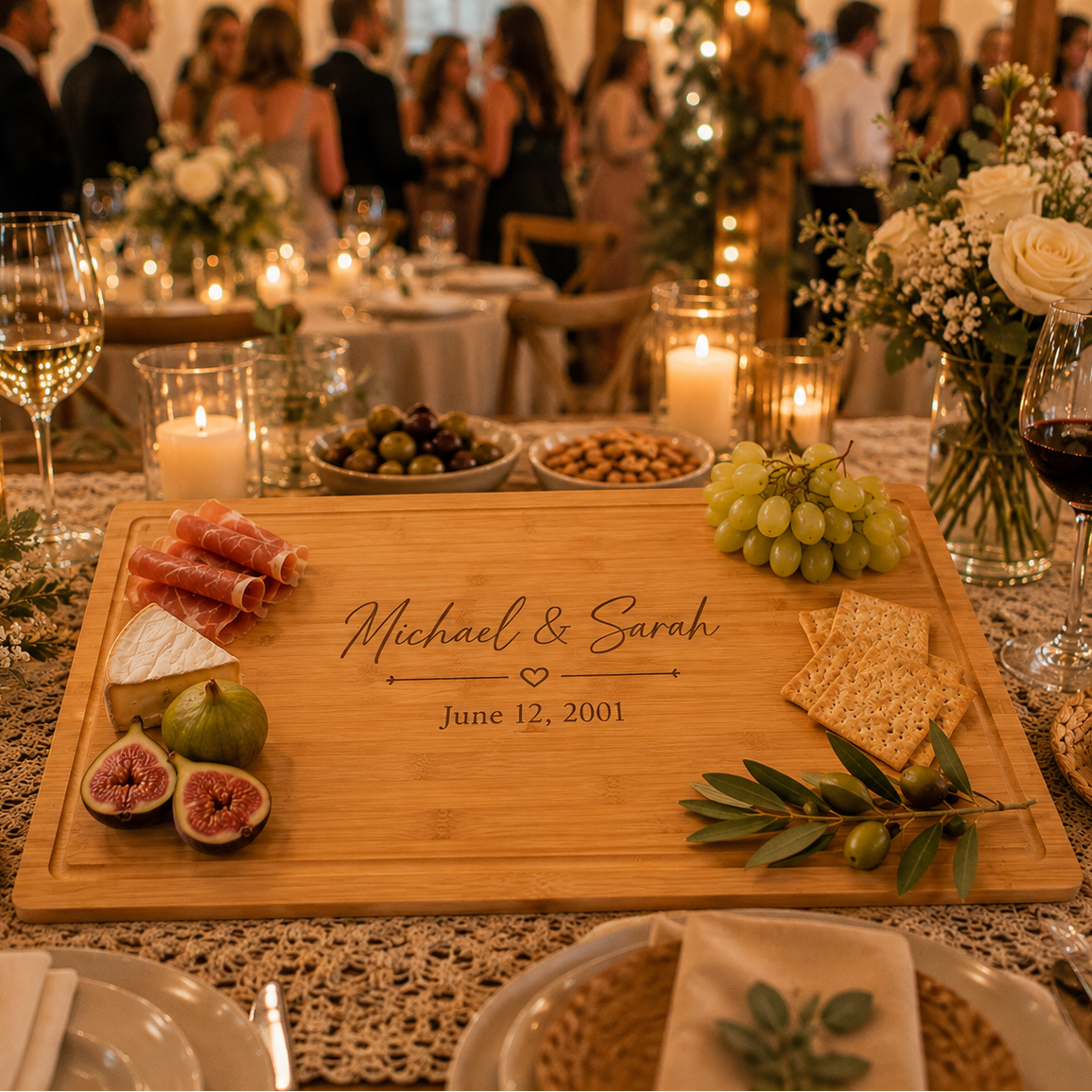 A personalized wooden charcuterie board engraved with 'Michael &amp; Sarah' and a date, styled on a decorated wedding reception table with cheese, grapes, crackers, olives, and candles in the background.