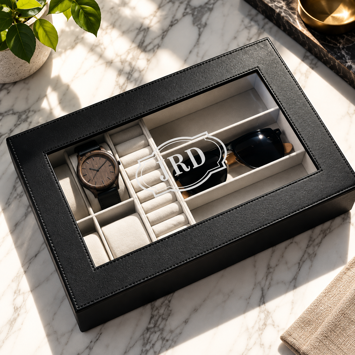 A black faux-leather watch and accessory organizer box with a clear top window, shown on a white marble surface. Inside the box are multiple compartments, including a watch, sunglasses, and a black item with a white monogram logo.