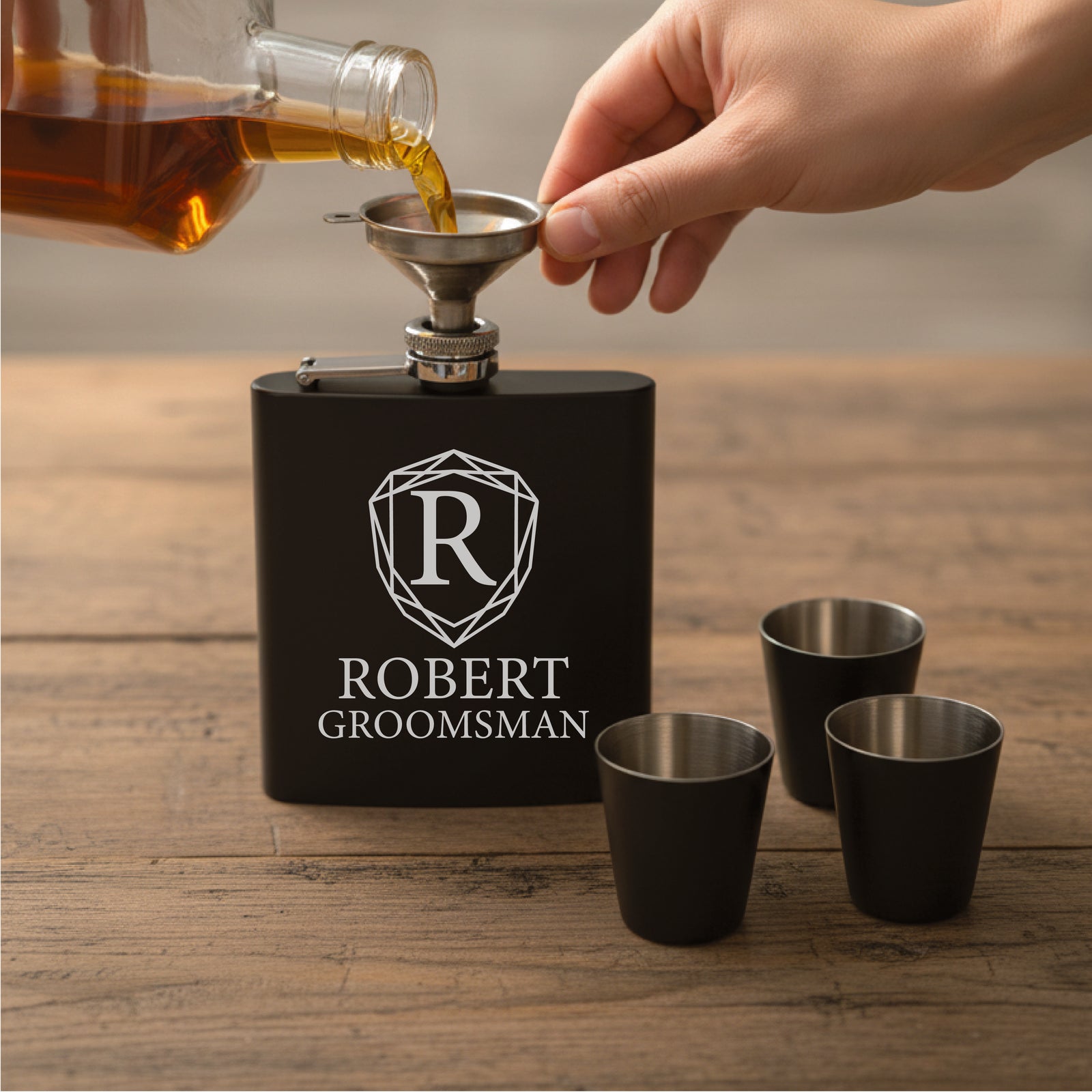 Black flask with stainless steel funnel pouring whiskey into matching shot glass, engraved with sample design showing "R" shield emblem and "ROBERT GROOMSMAN" text, three additional black shot glasses displayed beside it on wooden surface.