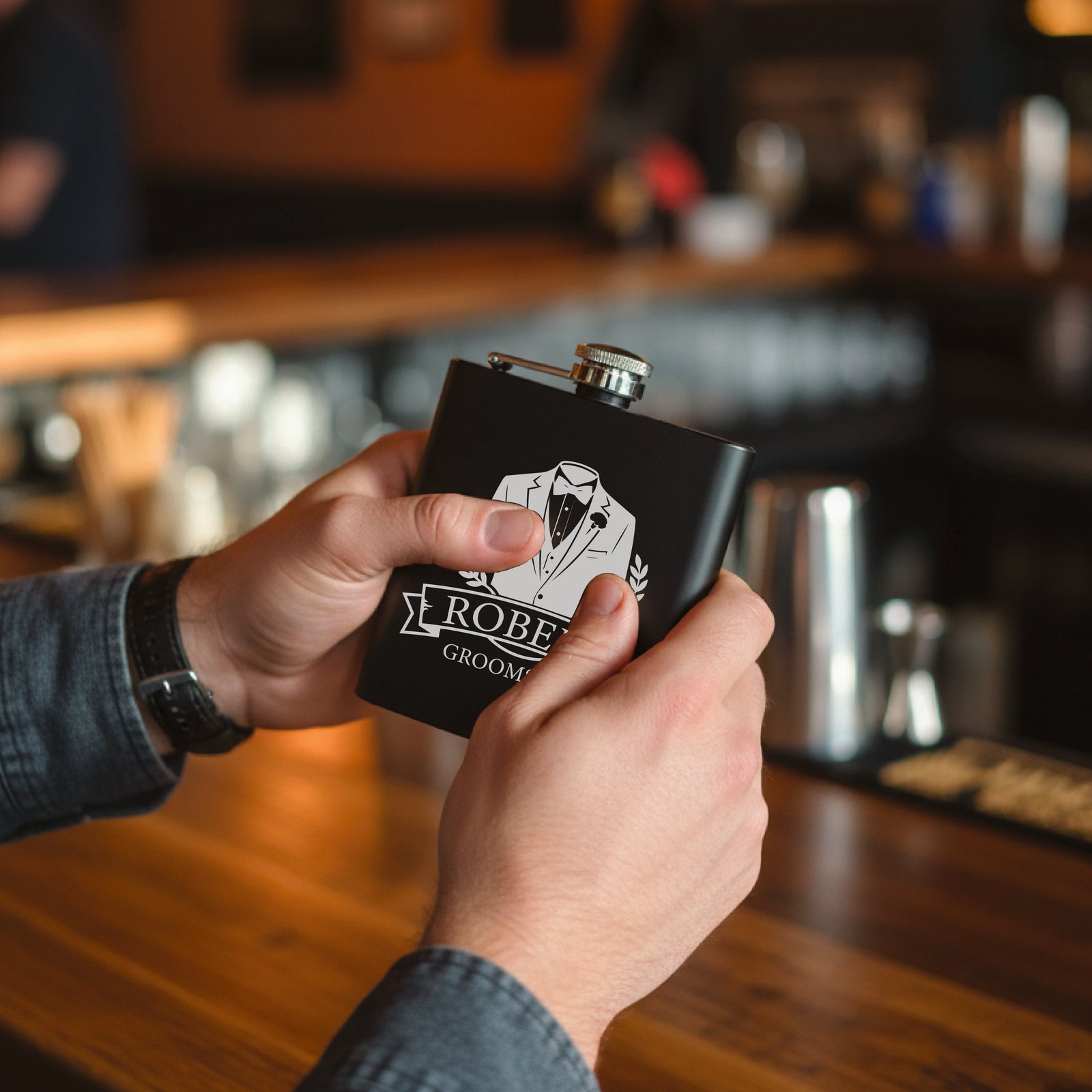 Black flask with stainless steel cap held in hands, engraved with sample design showing tuxedo graphic and "ROBERT GROOMSMAN" text, bar setting background