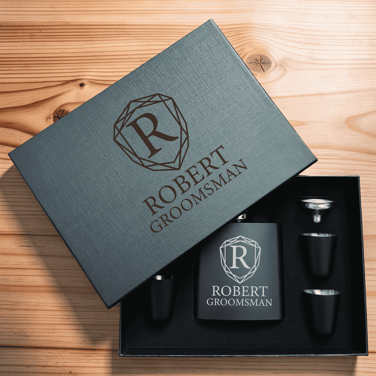 Gray linen gift box with geometric shield monogram and "ROBERT GROOMSMAN" engraving, opened to reveal black flask and shot glasses inside on wood surface