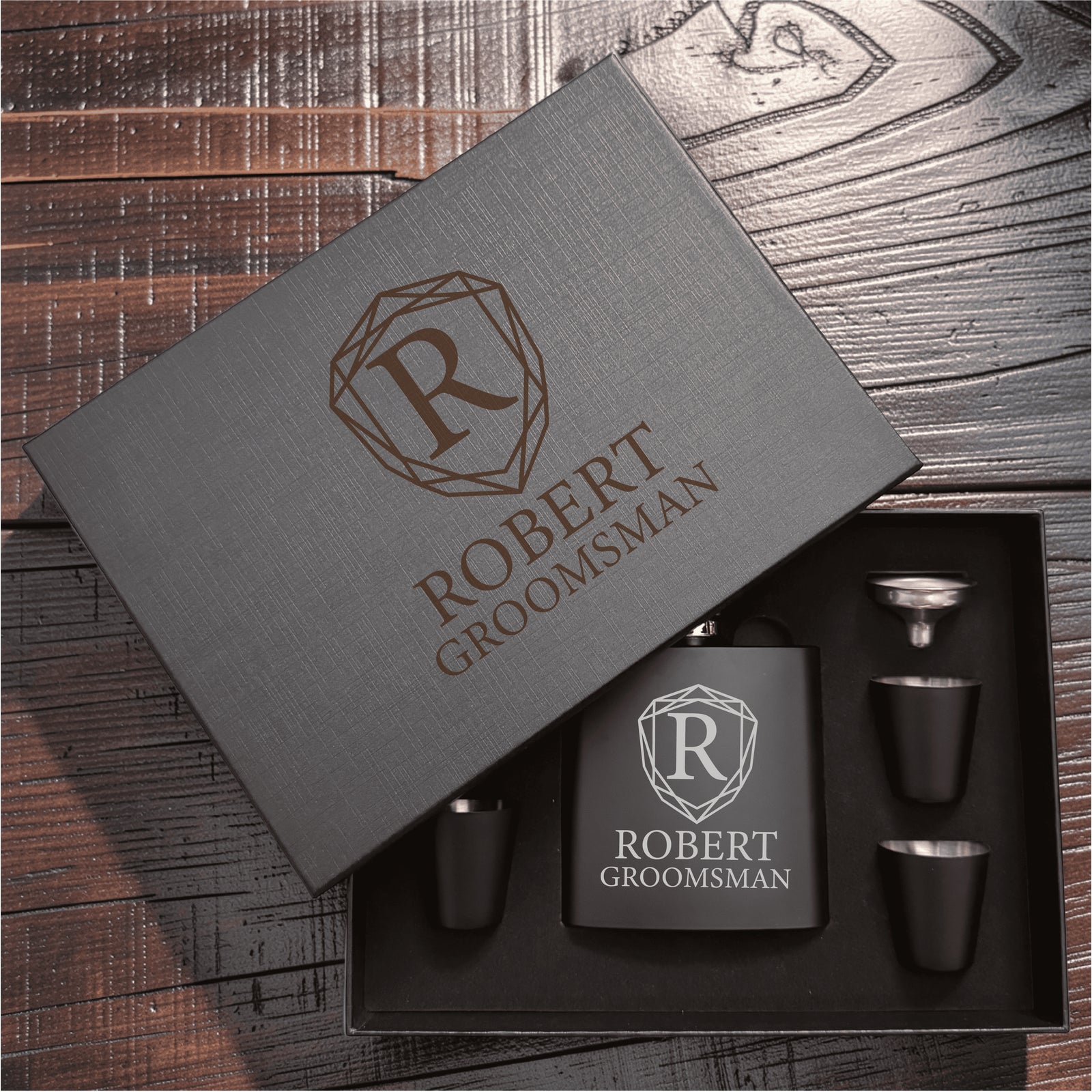 Gray gift box with personalized engraving reading "ROBERT GROOMSMAN" and geometric shield logo, containing black flask with matching shot glasses and funnel