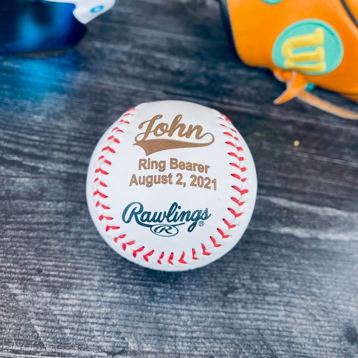 Engraved baseball groomsmen gift reading John, Ring Bearer and August 2, 2021 in warm brown on a white ball, set on dark wood with a baseball glove behind it
