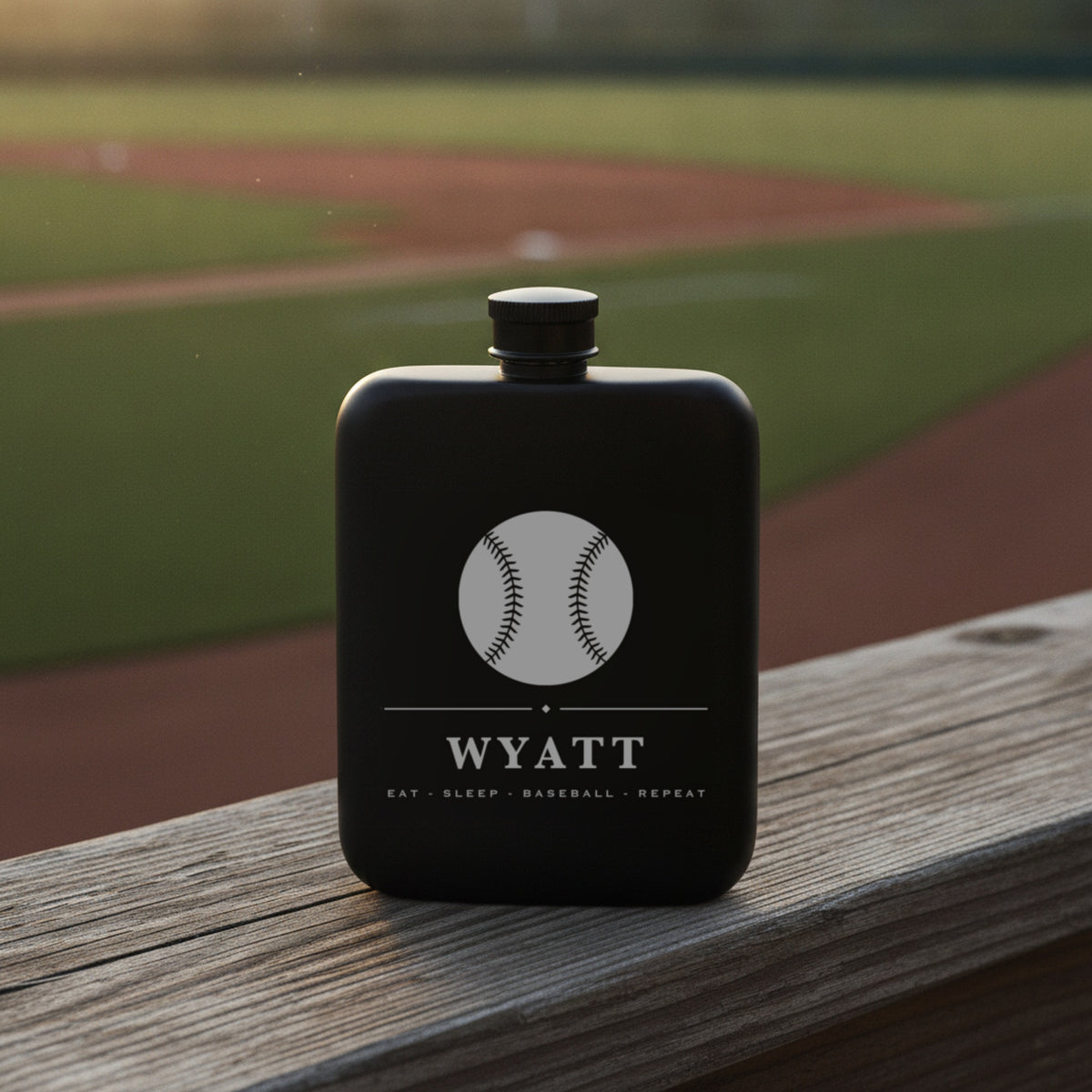 Engraved baseball groomsmen flask reading Eat Sleep Baseball Repeat on a weathered rail beside a green outfield in evening light.