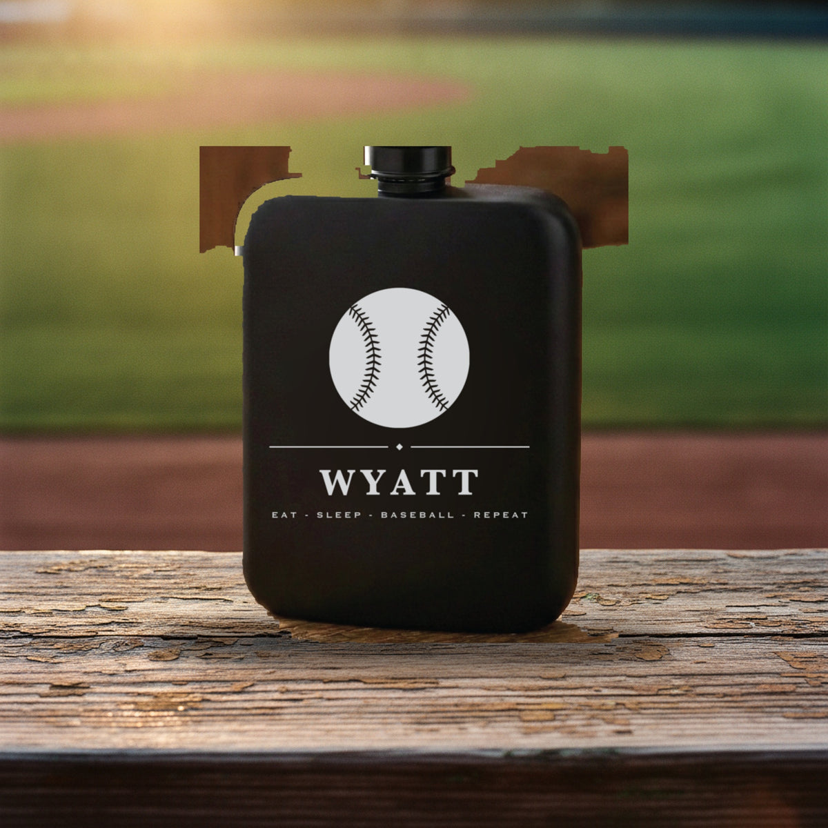 Engraved baseball groomsmen flask reading Eat Sleep Baseball Repeat on a weathered rail beside a green outfield in evening light.