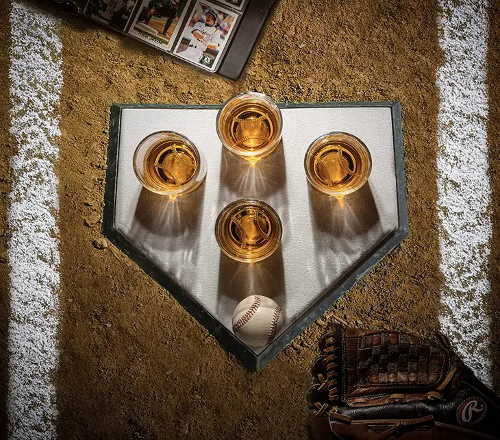 Baseball bat-shaped decanter set displayed on home plate-shaped stand with four whiskey glasses filled with amber liquid and baseball prop on infield dirt setting