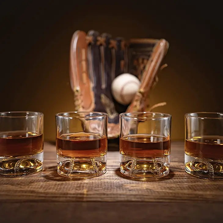 Four whiskey glasses filled with amber liquid arranged on wooden surface, with wooden baseball bat-shaped decanter and baseball glove with ball in soft-focus background.